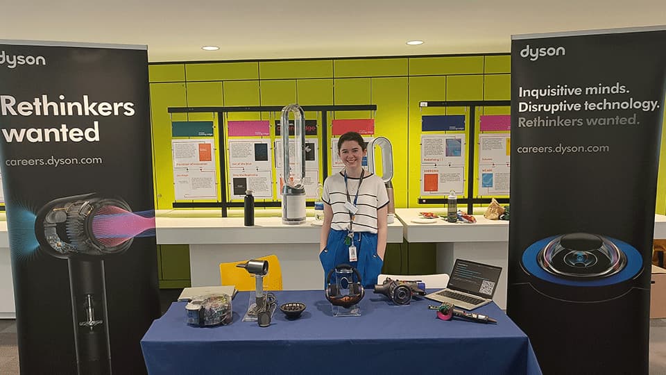 Georgia standing behind a table with a blue cloth. She is smiling and the banners behind her are promoting careers at Dyson.