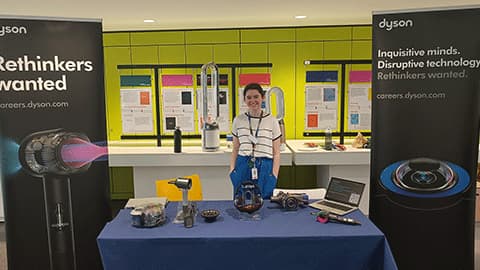 Georgia standing behind a table with a blue cloth. She is smiling and the banners behind her are promoting careers at Dyson.