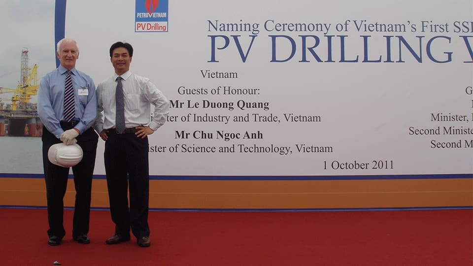 John in Singapore with a colleague when they named a new offshore drilling rig for PetroVietnam. John is wearing a blue shirt with a stripey tie and the other person is wearing a white shirt and black tie. They are standing in front of a sign that says naming ceremony of Vietnam's first SSI PV Drilling. Guests of Honour: Mr Le Duong Quang. 1 October 2011.