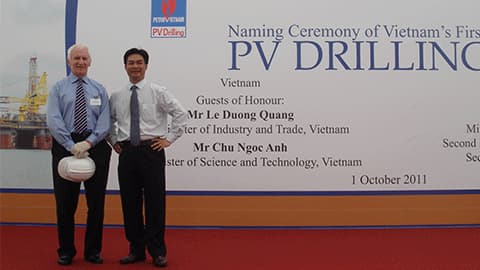 John in Singapore with a colleague when they named a new offshore drilling rig for PetroVietnam. John is wearing a blue shirt with a stripey tie and the other person is wearing a white shirt and black tie. They are standing in front of a sign that says naming ceremony of Vietnam's first SSI PV Drilling. Guests of Honour: Mr Le Duong Quang. 1 October 2011.