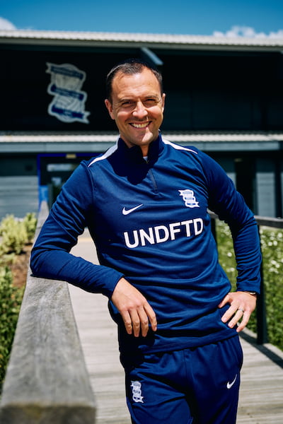 Chris wearing Birmingham City Football Club branded clothes in a dark blue colour, He leans on a wall, smiling.