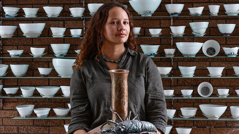 The background is a wall of 6 shelves with ceramic bowls and sculptures on it. Megan White is looking at the camera and holding one of her ceramic pieces which resembles a lobster on top of ceramic pot.