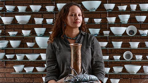 The background is a wall of 6 shelves with ceramic bowls and sculptures on it. Megan White is looking at the camera and holding one of her ceramic pieces which resembles a lobster on top of ceramic pot.