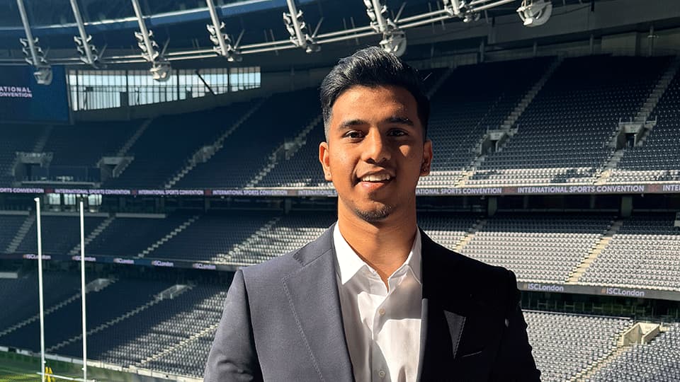 Vipul standing in front of a football pitch in a grey suit and white shirt. He is smiling at the camera in the sunshine.