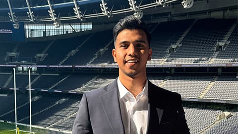 Vipul standing in front of a football pitch in a grey suit and white shirt. He is smiling at the camera in the sunshine.
