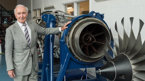 Desmond is wearing a grey suit and he is leaning on an engine.