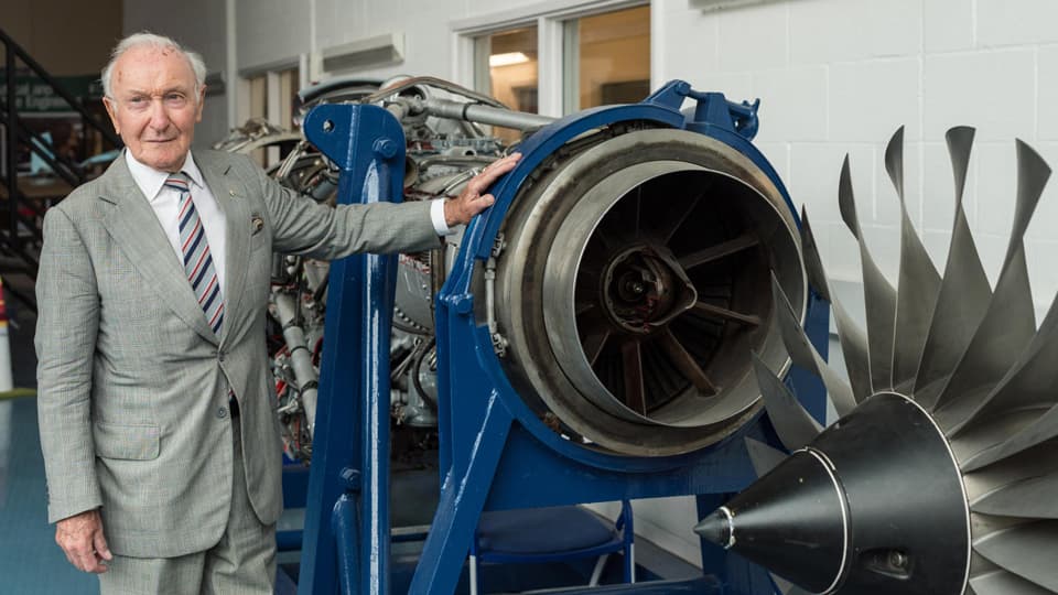 Desmond is wearing a grey suit and he is leaning on an engine.
