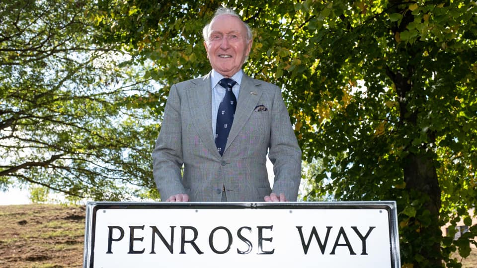 Desmond at the unveiling of Penrose Way in 2022. He is standing in behind the Penrose Way sign in Loughborough and he is wearing a grey suit.