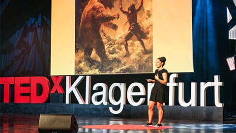 Elesa Zehndorfer wearing a black dress with her hair tied up and she is standing on stage talking at TEDx Klagenfurt, Austria.