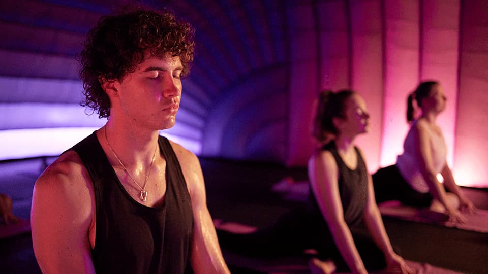 A man and two women doing the cobra pose in a Hotpod Yoga studio. There are dim lights which are pink and purple around the edges of the room. They all have there eyes closed and look relaxed.