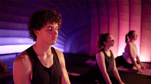 A man and two women doing the cobra pose in a Hotpod Yoga studio. There are dim lights which are pink and purple around the edges of the room. They all have there eyes closed and look relaxed.