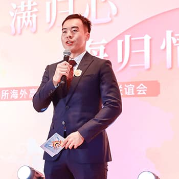 Winston is wearing a black suit with a pocket chief. He is holding a microphone and speaking and a bit of card looking out at an audience.