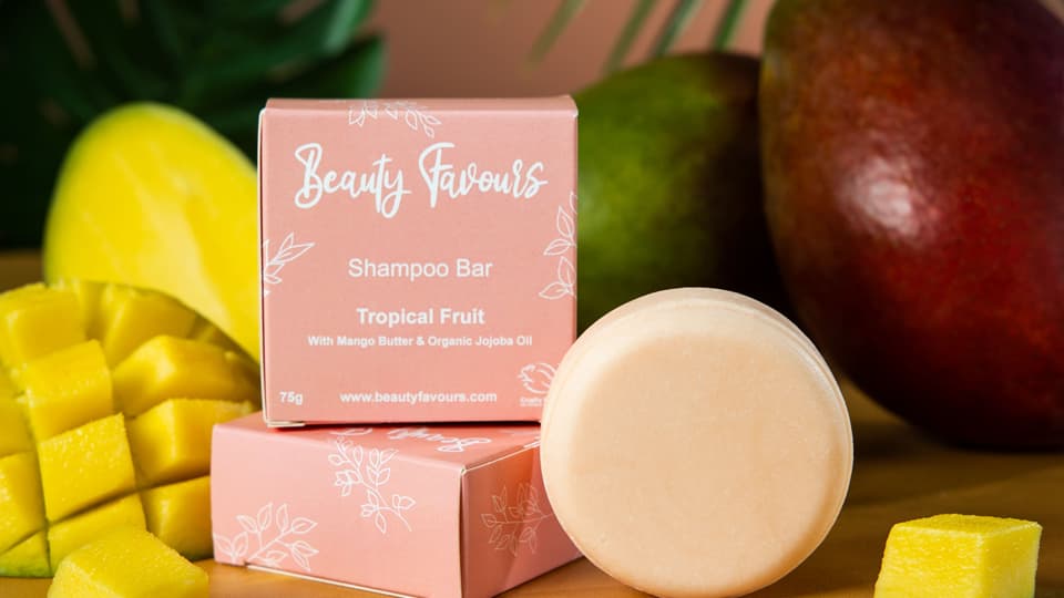 A round pink shampoo bar and a square, pink package for the shampoo bar. There are mangoes in the background.