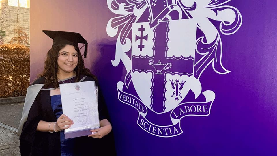 Alumna Liliane Santos is wearing a graduation gown and cap. She is holding up her degree certificate and is standing in front of a banner which is purple and has the University's logo and motto in white text Veritate, Scientia, Labore.
