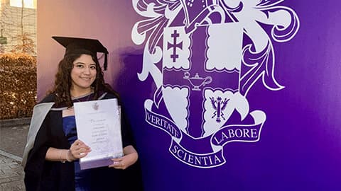 Alumna Liliane Santos is wearing a graduation gown and cap. She is holding up her degree certificate and is standing in front of a banner which is purple and has the University's logo and motto in white text Veritate, Scientia, Labore.