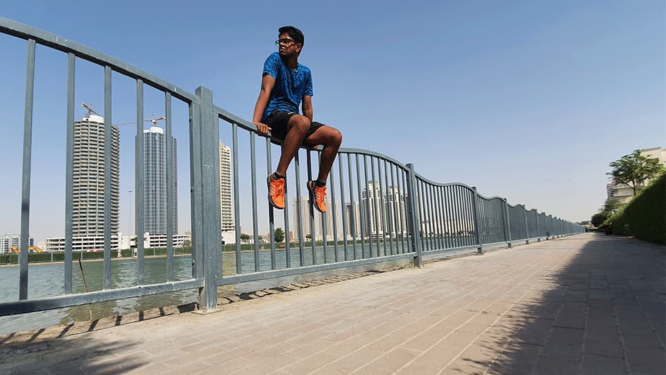 Mridul is in Dubai sitting on a fence in his running gear with blue sky and the sun is shining. There are tall buildings in the background.