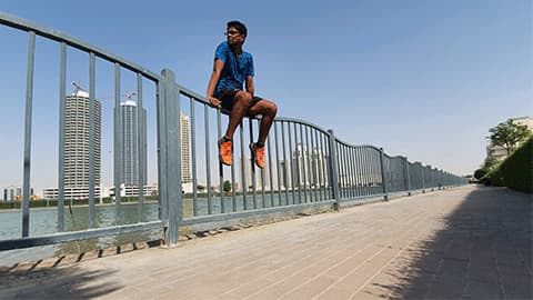 Mridul is in Dubai sitting on a fence in his running gear with blue sky and the sun is shining. There are tall buildings in the background.
