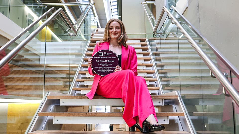 Alumna Sri is holding the Purple Plaque with her name on it on Loughborough Campus. She is smiling and holding the award up and she is wearing a bright pink suit while sitting on some stairs.