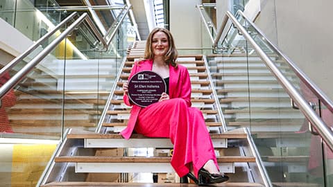 Alumna Sri is holding the Purple Plaque with her name on it on Loughborough Campus. She is smiling and holding the award up and she is wearing a bright pink suit while sitting on some stairs.