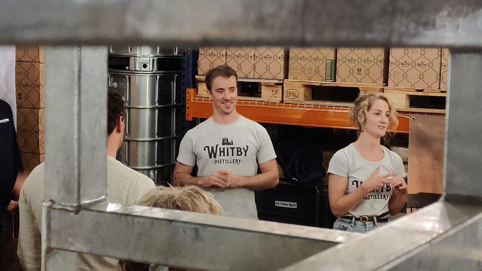 Alumni Jessica and Luke wearing white t-shirts which read Whitby Distillery on them. They are working and smiling in Whitby Distillery together. The image is frame by some metal equipment and they are the focal point.