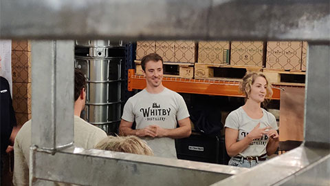 Alumni Jessica and Luke wearing white t-shirts which read Whitby Distillery on them. They are working and smiling in Whitby Distillery together. The image is frame by some metal equipment and they are the focal point.
