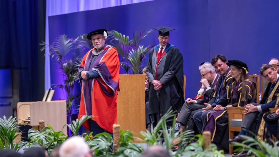 Sir Michael Berry standing on stage wearing a cap and gown at graduation