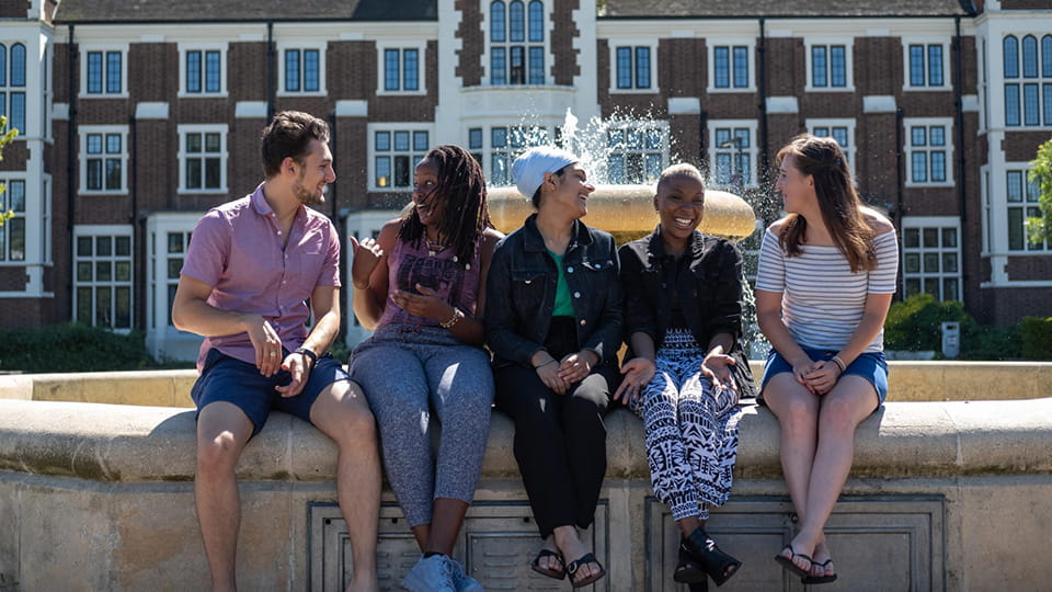 Five students sitting in a row with a fountain and historic university building in the background