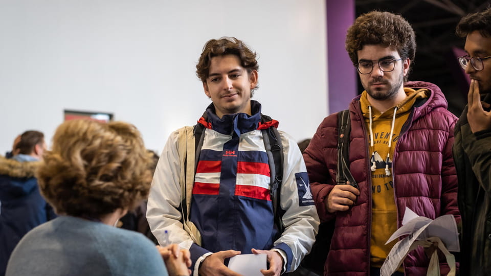 Three students stand facing towards a person who is seated. The students are wearing coats.