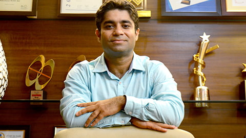 Aman Dhall with his arms folded leaning on something. There are trophies on shelves in the background.
