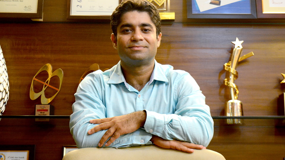 Aman Dhall with his arms folded leaning on something. There are trophies on shelves in the background.