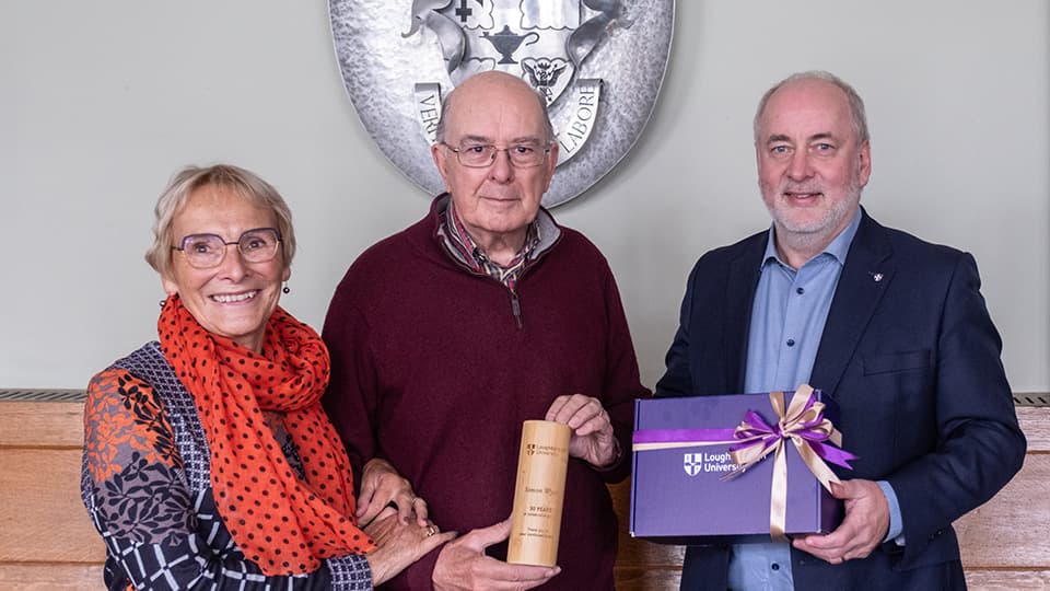 Three people stand together smiling. Simon, centre, holds a commemorative plaque.