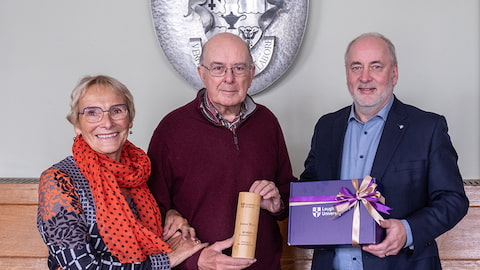 Three people stand together smiling. Simon, centre, holds a commemorative plaque.