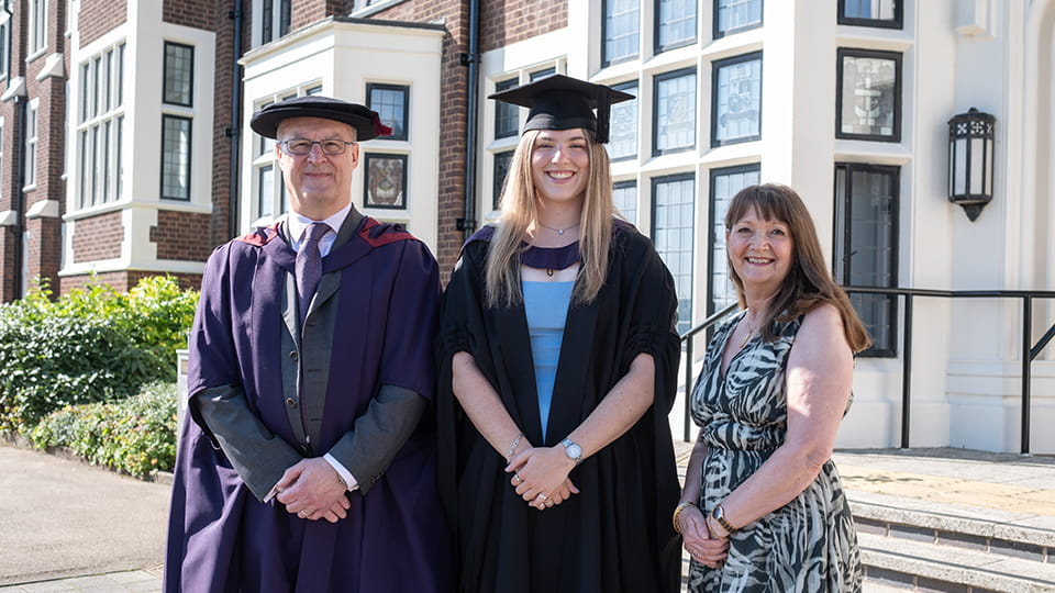 Three people standing outside Hazlerigg Building. Two of them wear graduation gowns and hats.