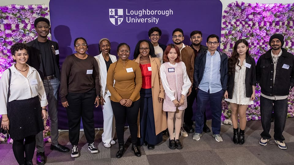 A group of Global Impact Scholars standing in front of an African Violet Loughborough University banner and two flower walls