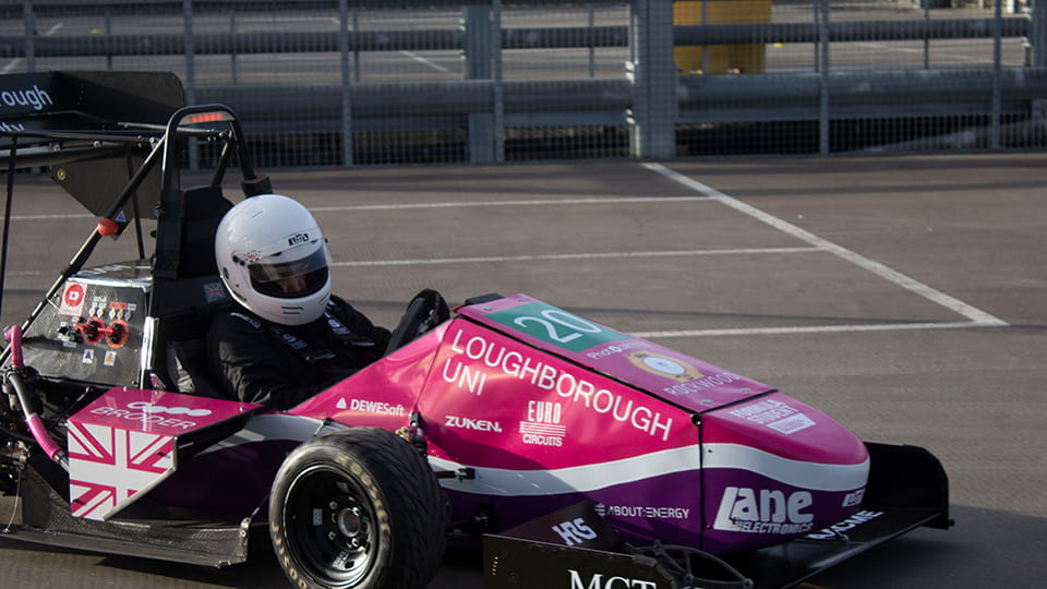 A driver in the LUMotorsport car in a car park. The car is predominantly pink and has a variety of logos on it.