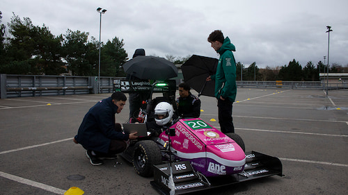 A driver in the LUMotorsport car in a car park. The car is predominantly pink and has a variety of logos on it. There are people with umbrellas around the car and one holds a laptop.