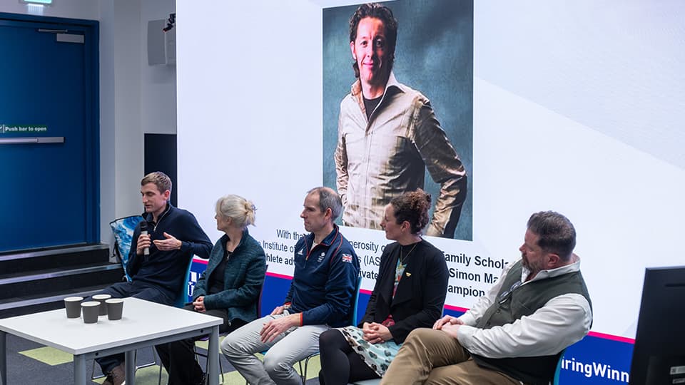 Four people seated in a panel style, one holds a microphone. A photo of Dr Simon Marshall is on a screen behind them.