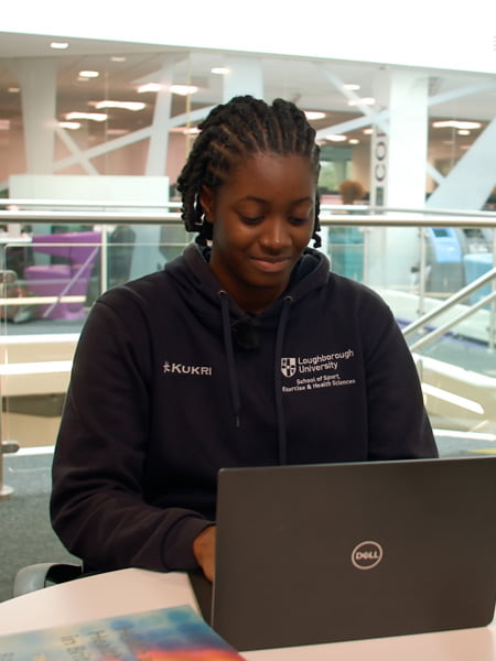 Student, Joy, is seated in the library, smiling looking at a laptop
