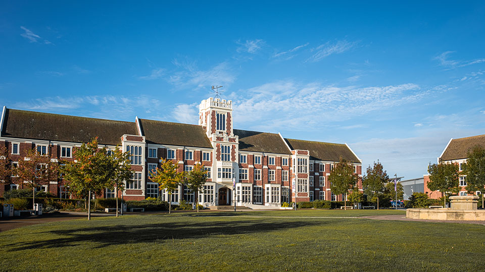 A low-angle shot of a brick building with a tower, set against a blue sky. Red-leaved trees flank a paved walkway in front.