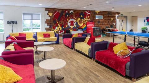 A common room with colourful sofas, round tables, a ping pong table, and a mural on the back wall. There are windows, overhead lights, and various recreational items visible.