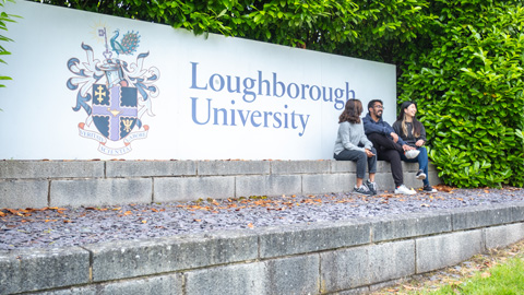 Three people sit on a low wall in front of a Loughborough University sign bordered by green foliage.