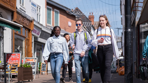 A group of students walk down a sunny street lined with shops and cafes, some carrying backpacks and chatting together.