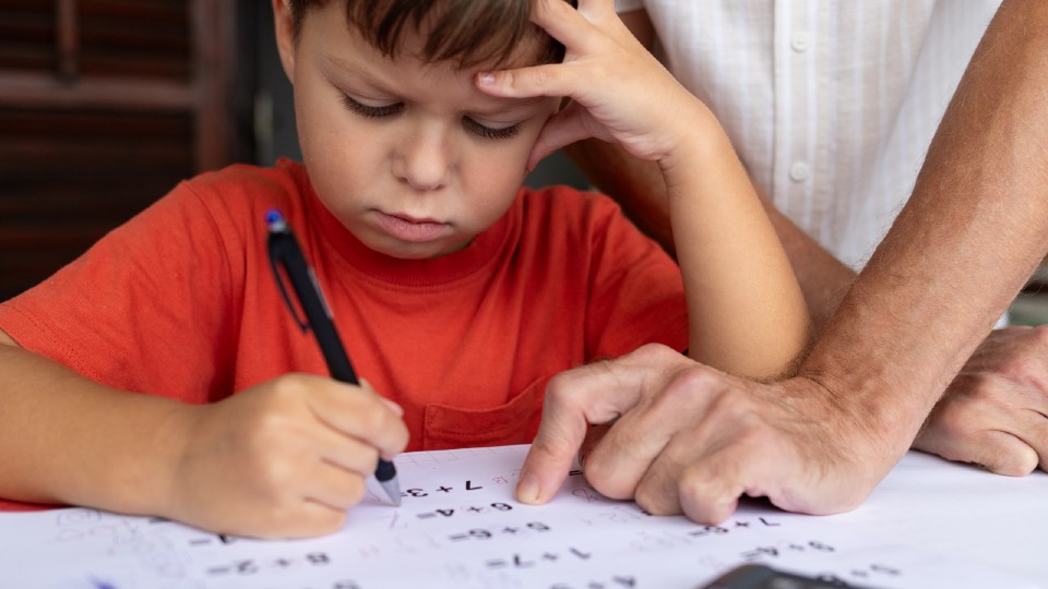 A young boy in a red t shirt holding a pen doing his math homework while an adult, out of shot, points at the numbers on the homework sheet