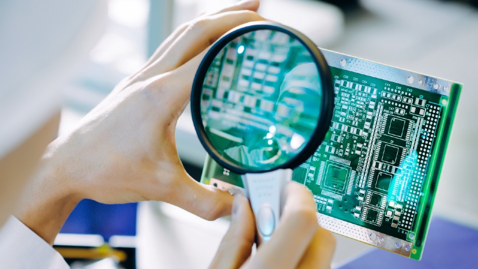 A person holding a magnifying glass to a microchip board.
