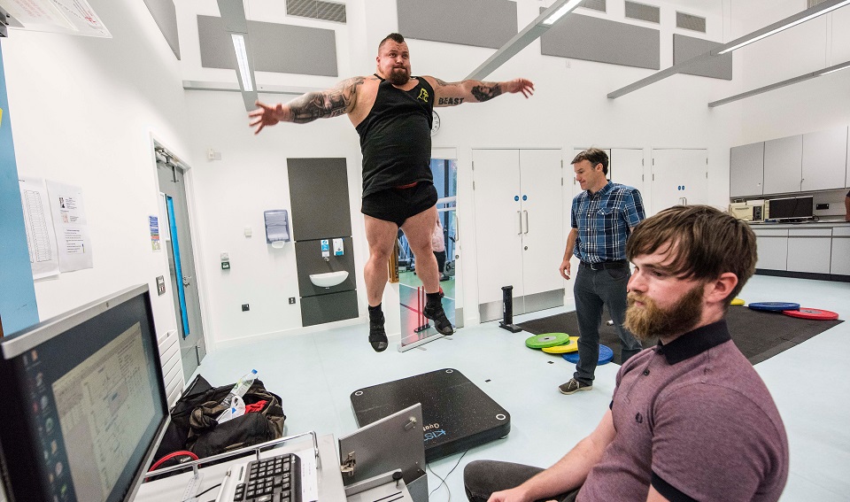 Strongman Eddie Hall in a science lab carrying out a jumping test with two academics.