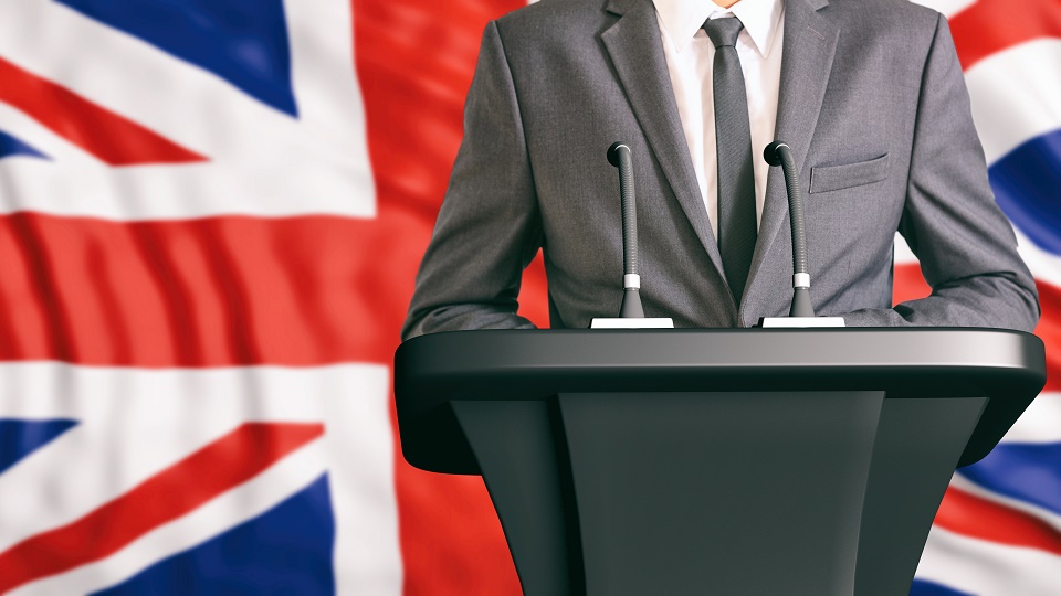 A male politician in a suit is stood at a lecturn in front of the Union Jack flag