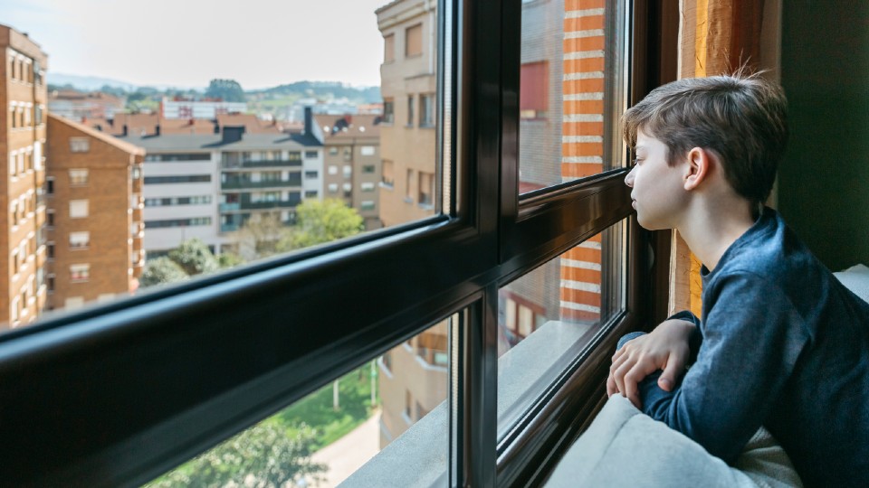A teenage boy sat on a bed looking out the window and a cityscape.