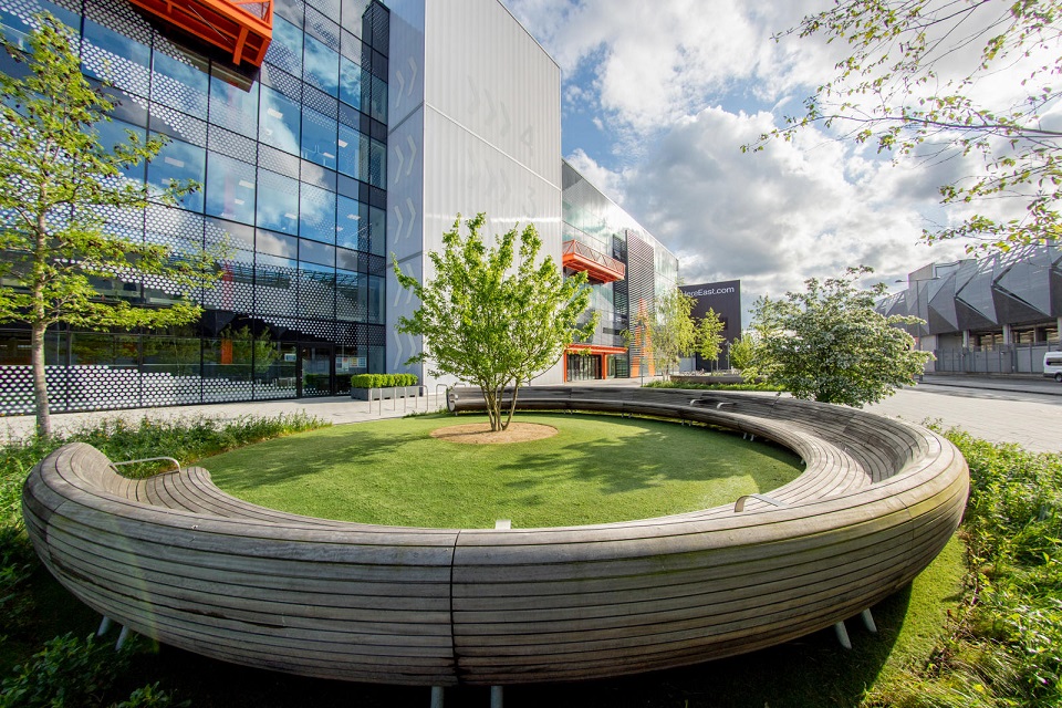 An outside shot of the Loughborough University London campus, woith a circular seating area, trees and the building in the background.