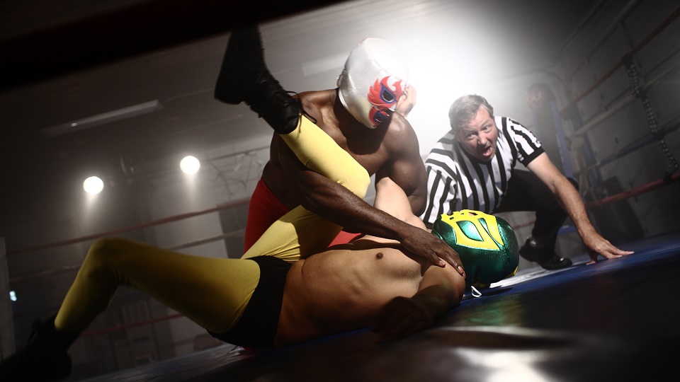 Image: Two wrestlers are fighting in a boxing ring wearing head masks, with a referee knelt beside them.