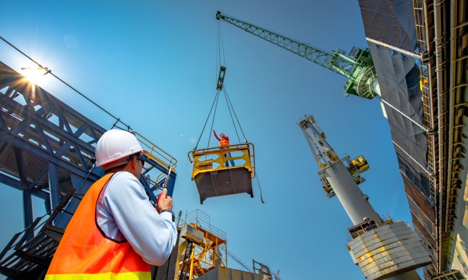 A man in a hard hard and high-vis vest is stood on a construction site looking up at a crane lifting goods